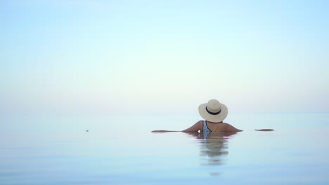 Zurück-Zur-Kamera-Eine-Junge-Frau-In-Einem-Infinity-Pool-Eines-Resorts,-Der-Auf-Einen-Endlosen-Blick-Auf-Das-Meer-Hinausblickt