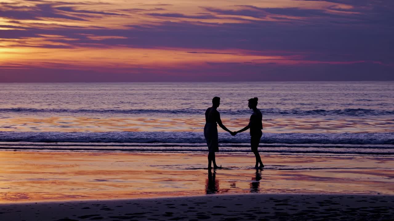 Silhouetted couple holding hands on a beach at sunset, captured from a low angle