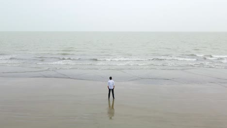 Hombre-De-Pie-En-La-Orilla-Del-Mar-Frente-A-La-Cámara-Mirando-El-Vasto-Océano-Índico-Vestido-Con-Mangas-Largas-Blancas-Y-Jeans-Con-Las-Manos-En-El-Bolsillo