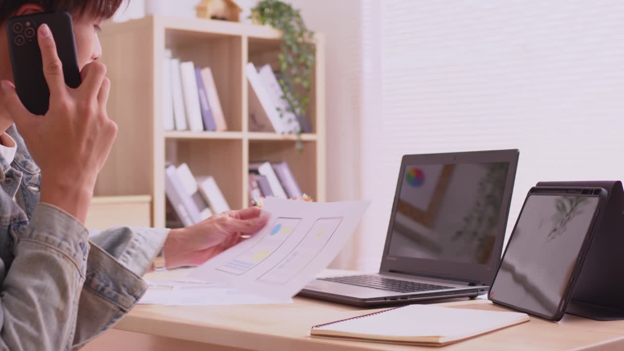 el diseñador asiático usa su teléfono inteligente para comunicarse, trabajar en proyectos y papeles, y trabajar desde casa en un escritorio con una computadora portátil.