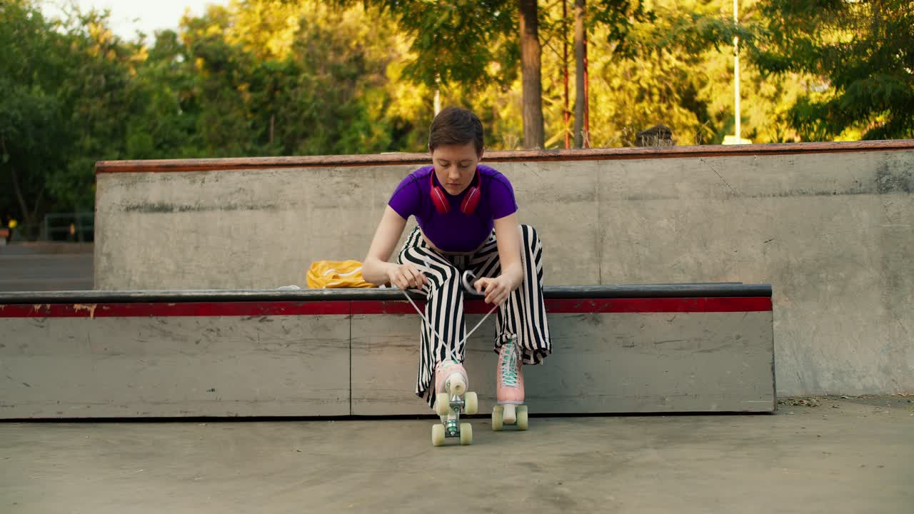 Shooting from the front: A young short-haired girl in a purple top and striped pants lace up her skates on a special bench in a skate park for skaters
