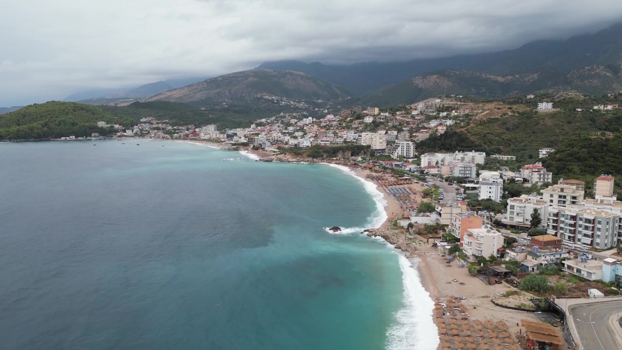Veduta aerea con drone del litorale della spiaggia di Faliraki a Rodi, Grecia, che mostra una spiaggia sabbiosa curva, mare turchese, edifici costieri, ombrelloni da spiaggia e colline verdi sotto cieli nuvolosi.