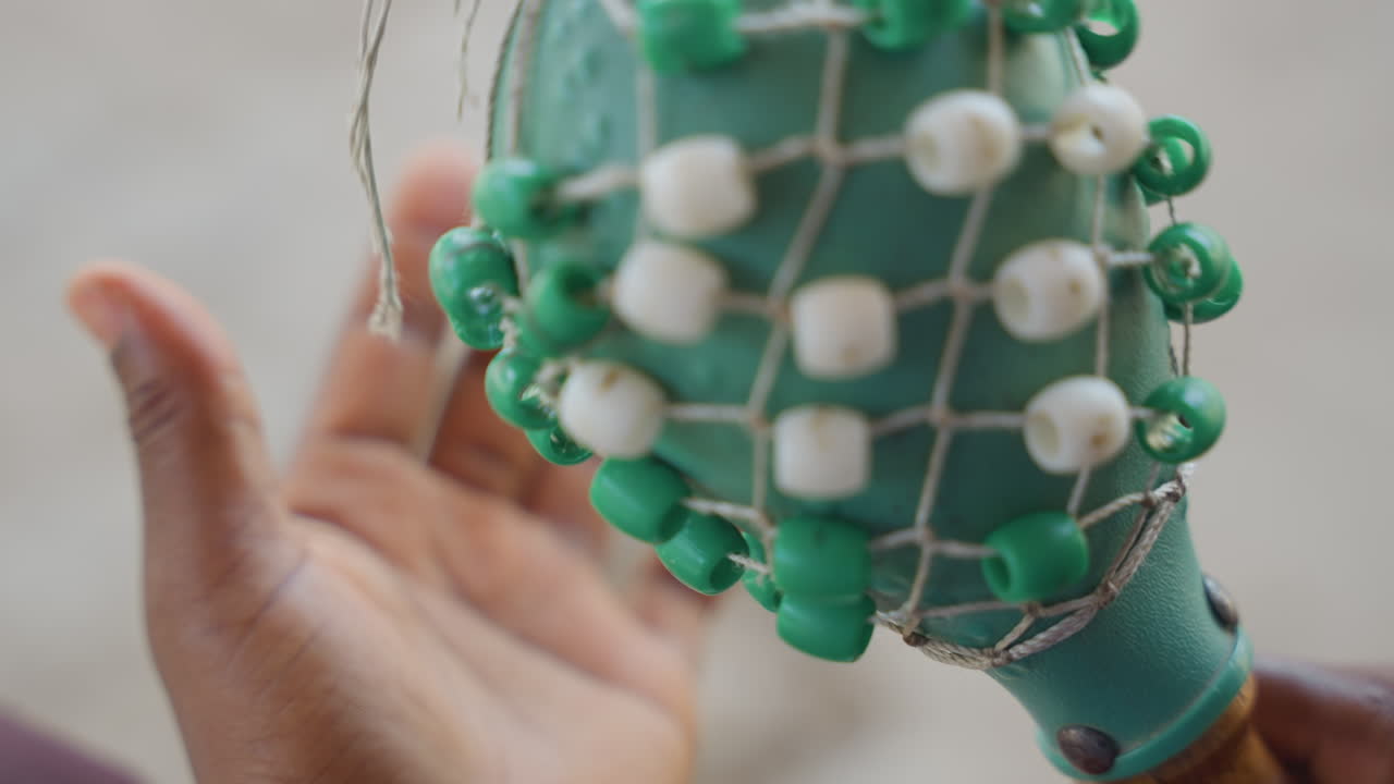 Nahaufnahme von schwarzen Händen, die Perlen inspizieren. Detaillierte Ansicht eines grün-weißen Perlennetzes auf einem Shaker, mit texturiertem Netz und Quaste, sandiger Hintergrund, sorgfältige Untersuchung mit langsamen, gezielten Bewegungen, Fokus.