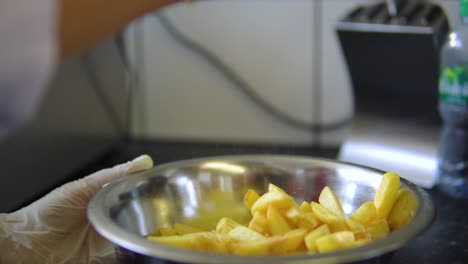 Fast-food-restaurant-worker-with-white-protective-gloves-on-is-salting-fried-chips-fries-in-a-bowl-slow-motion