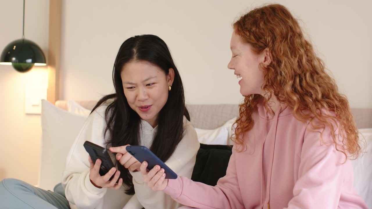 Amiche diverse che mostrano e si scambiano smartphone sul letto dopo aver ricevuto un'offerta telefonica, confrontando gli schermi
