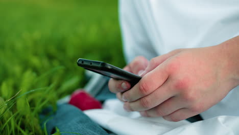 close-up of hand holding smartphone and operating it while seated on grassy field, red cloth partially visible beside the hand, surrounded by greenery