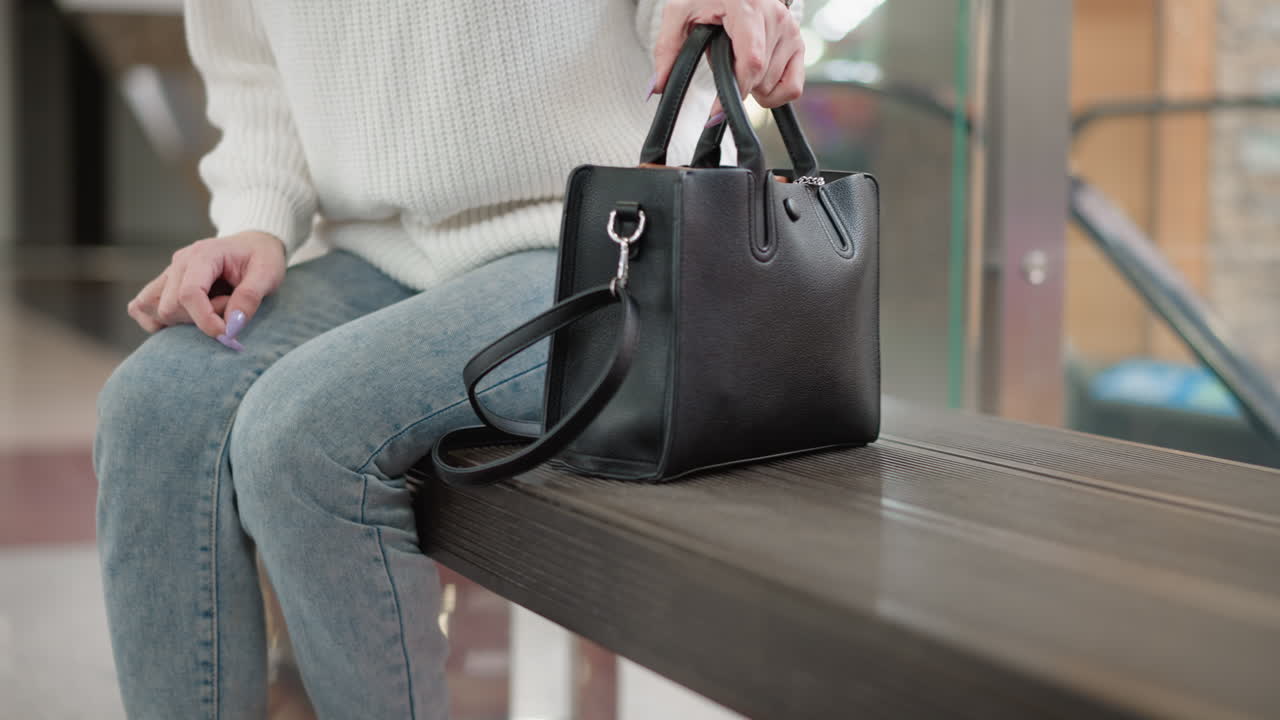 lower body framed shot of youthful woman in denim jeans and boots holding black leather bag, placing bag on bench beside moving walkway then sitting down, emphasis on bag texture and casual motion