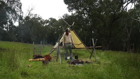 a bush settler cooks over a fire with a billy at a hisotorical pioneer camp