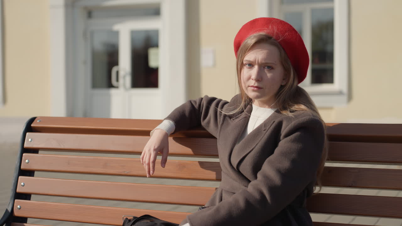 Giovane donna con basco rosso. Una donna siede con sicurezza su una panchina cittadina. La donna con il cappello rosso guarda serenamente la fotocamera. Una donna in stile streetwear seduta con sicurezza contro lo sfondo di una facciata cittadina.
