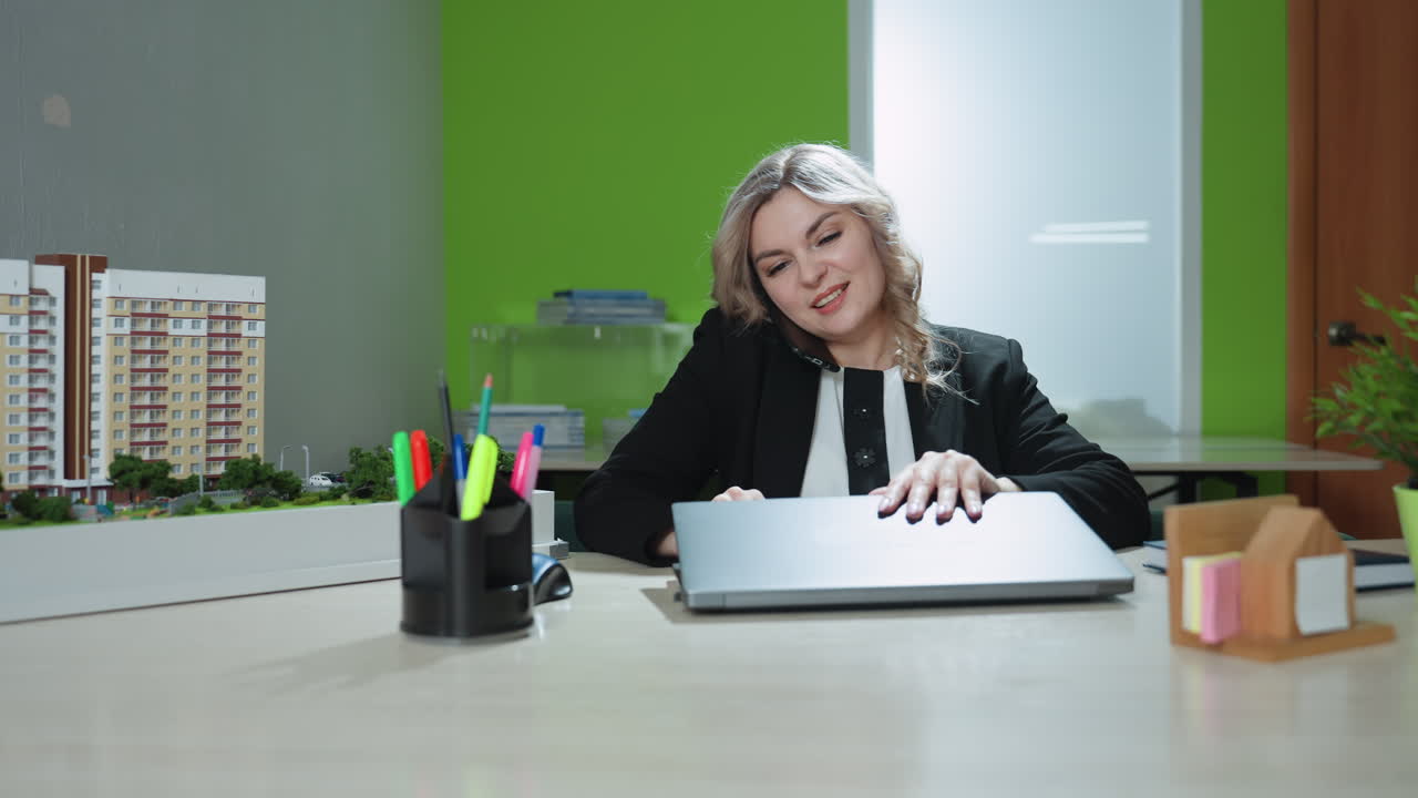 Real estate agent sits at office desk multitasking during warm phone call while opening laptop, surrounded by building model, documents, stationery, and potted plants in professional work environment