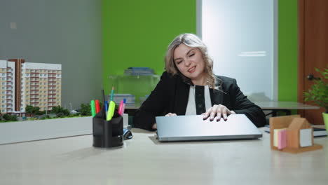 real estate agent sits at office desk multitasking during warm phone call while opening laptop, surrounded by building model, documents, stationery, and potted plants in professional work environment