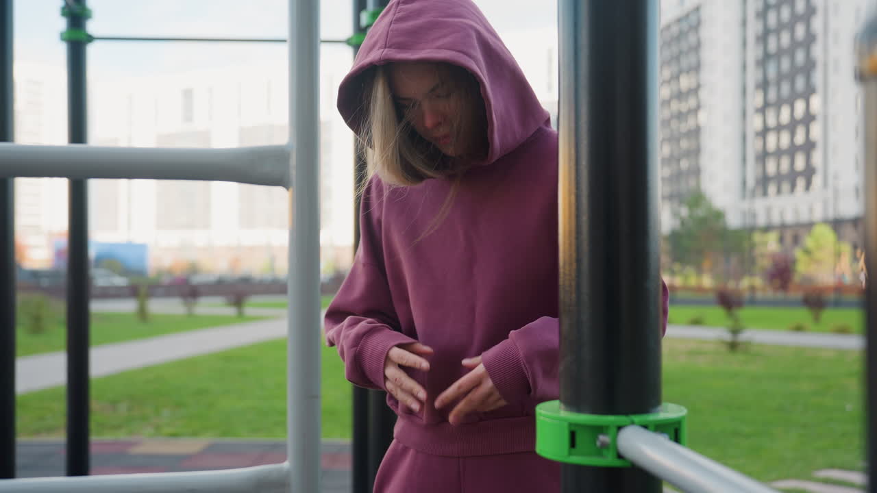 Outdoor trainer in hooded tracksuit holds sports bag and flips arms for wrist warmup before engaging in full body calisthenics among metal bars under open sky near modern apartments