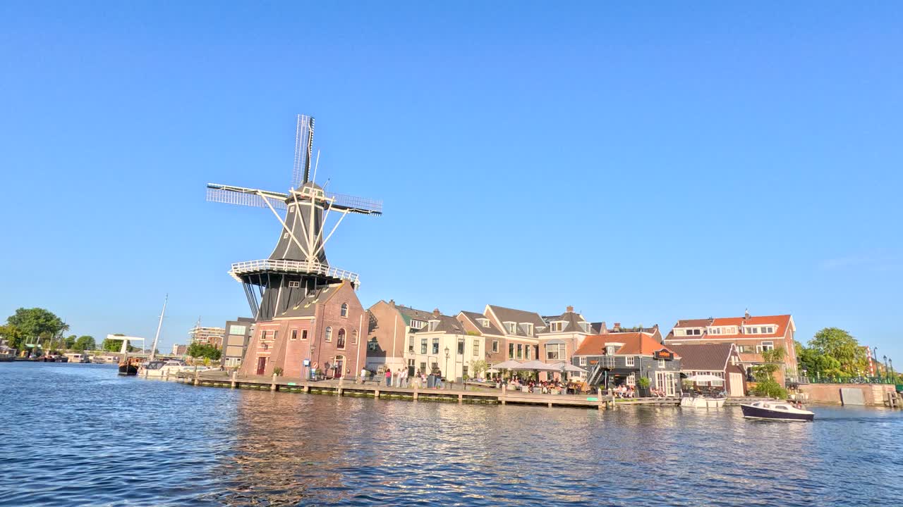 Sunny daytime boat ride along canal, passing windmill, historic buildings, and waterfront reflections