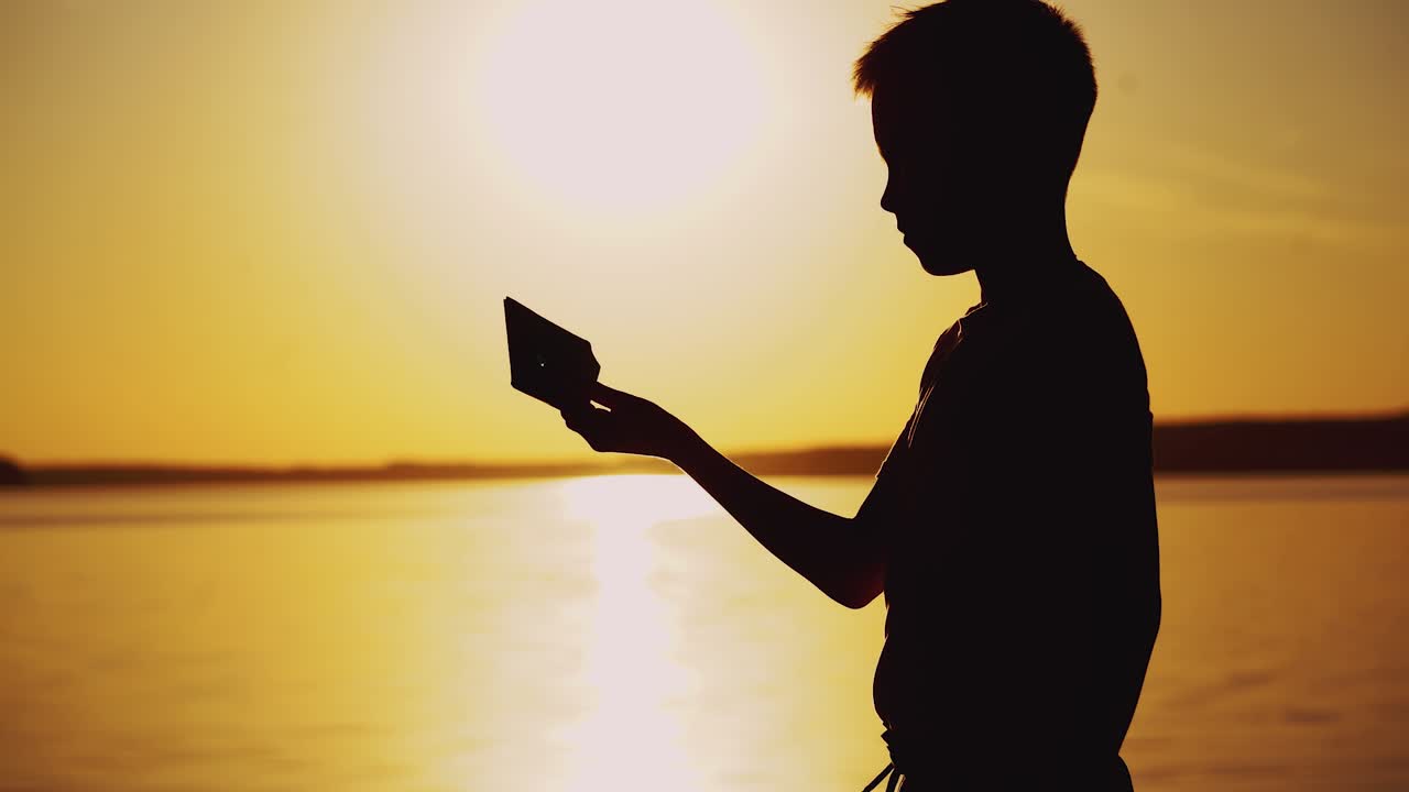 Little boy imitating swimming with his homemade boat near the river at sunset. Silhouette of a child with origami ship made from paper