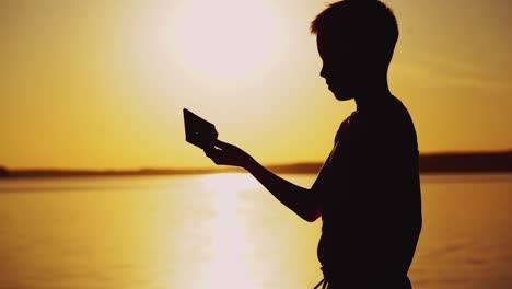 little boy imitating swimming with his homemade boat near the river at sunset. silhouette of a child with origami ship made from paper