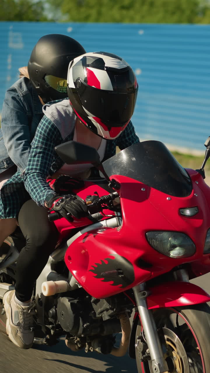 dos parientes montan una bicicleta eléctrica roja, ambos con cascos, están montando cerca de un lugar cubierto con láminas de zinc azul con un edificio y un coche estacionado dentro mientras pasan