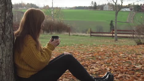 una chica adulta está sentada bajo el árbol y bebiendo una bebida caliente en el paisaje otoñal en cámara lenta