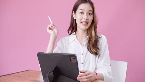 Feliz-Y-Exuberante-Mujer-Asiática-Vestida-De-Blanco-Sentada-En-Una-Silla-Blanca-Hablando-Lentamente-Mientras-Usa-Una-Tableta-Electrónica-Contra-Un-Fondo-Rosa-Aislado