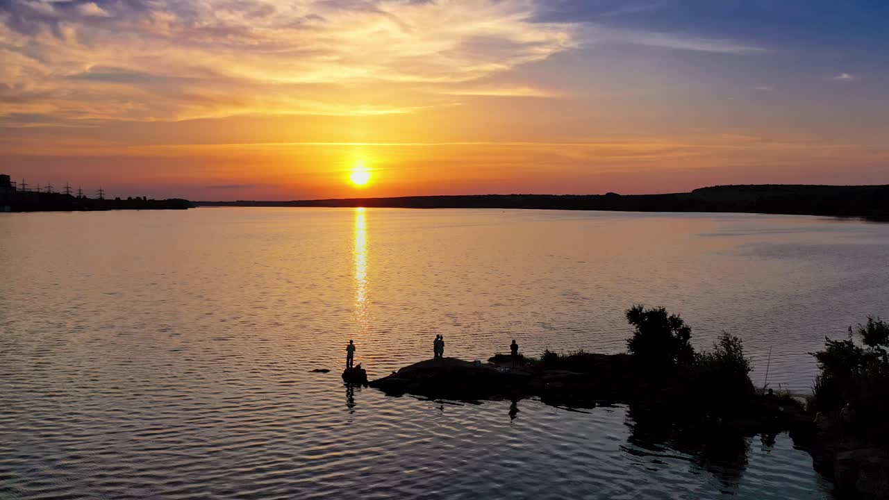 View from a drone on the river in the evening. People stand on long stones situated in water at sunset. Camera moves forward.