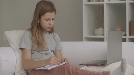 school girl is listening teacher by laptop and writing at exercise book sitting on couch at home e-learning for schoolers