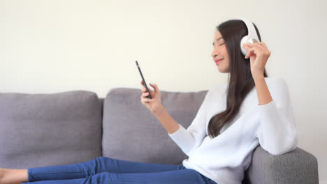 happy pretty asian woman watching phone and listen to music while lying on the couch wearing overhead headphones, smiling and snapping fingers to the beat, side view