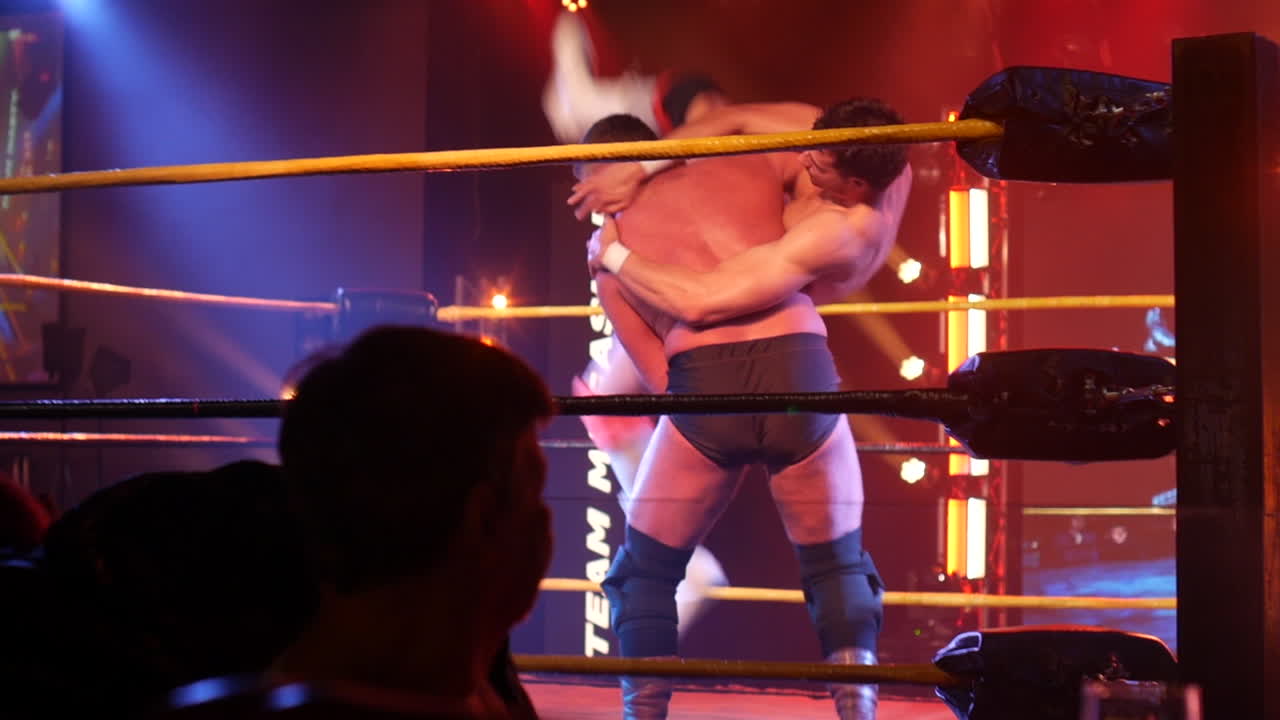 A wrestler in black trunks lifts his opponent high over his shoulders in a powerful maneuver inside the wrestling ring.