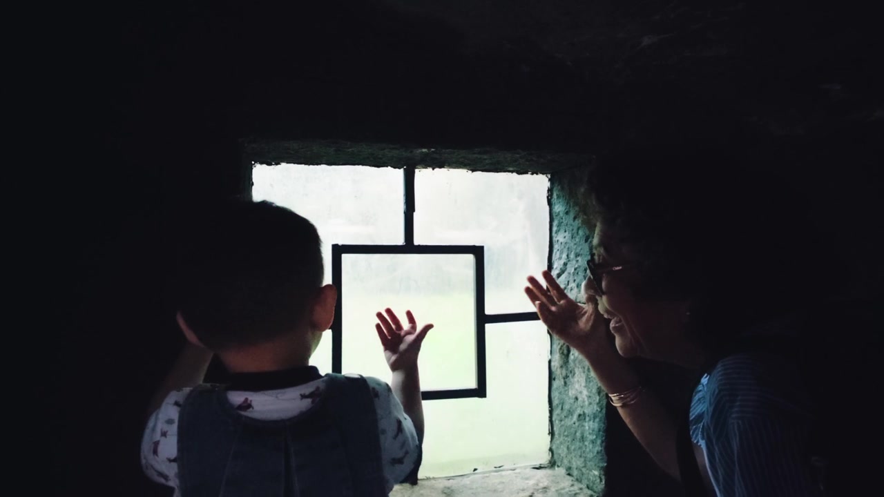 A Latin grandmother and a baby are seen inside a mammal habitat at Chapultepec Zoo, peering through an observation window and calling out, engaging with the wildlife.