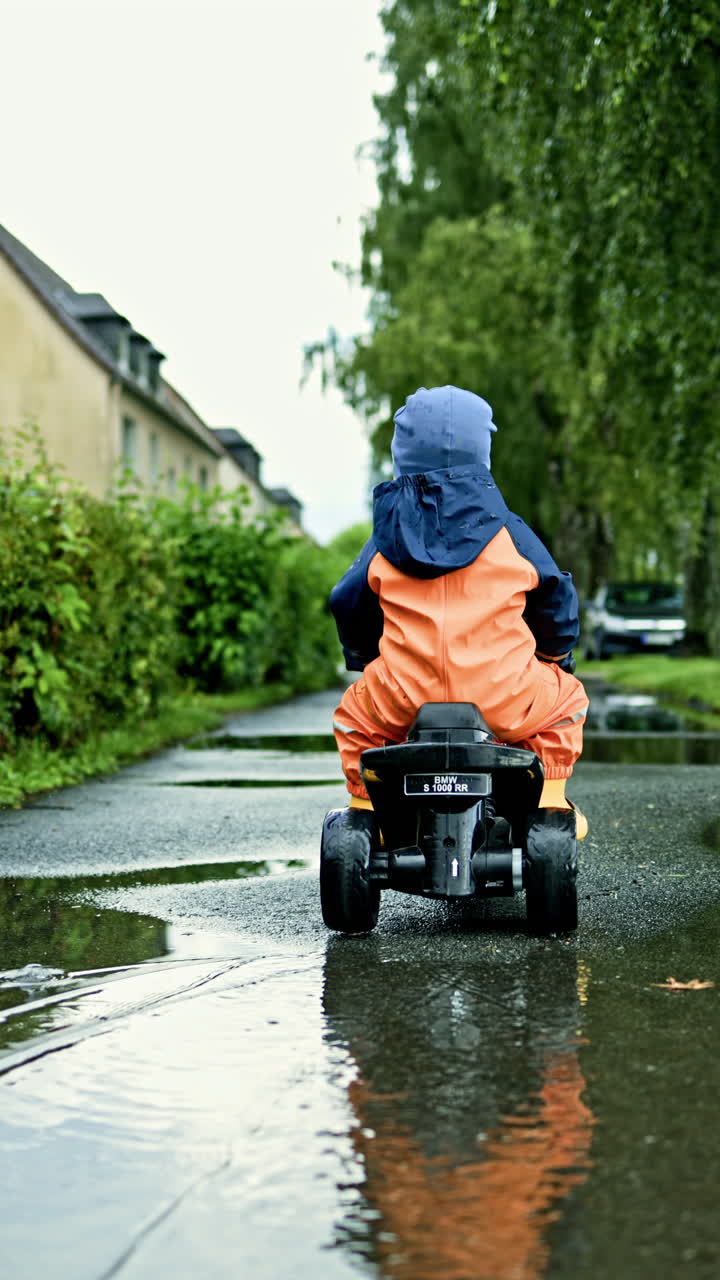 Toddler rides a children's electric motorcycle. A 2-year-old boy rides through puddles after the rain. A child in a waterproof overalls and rubber boots walks in the rain outside