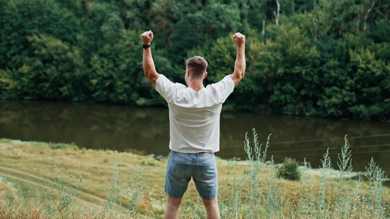 Rear view of a strong muscular man in white t-shirt and jeans shorts. Man raises hands rejoicing life and free time in nature.