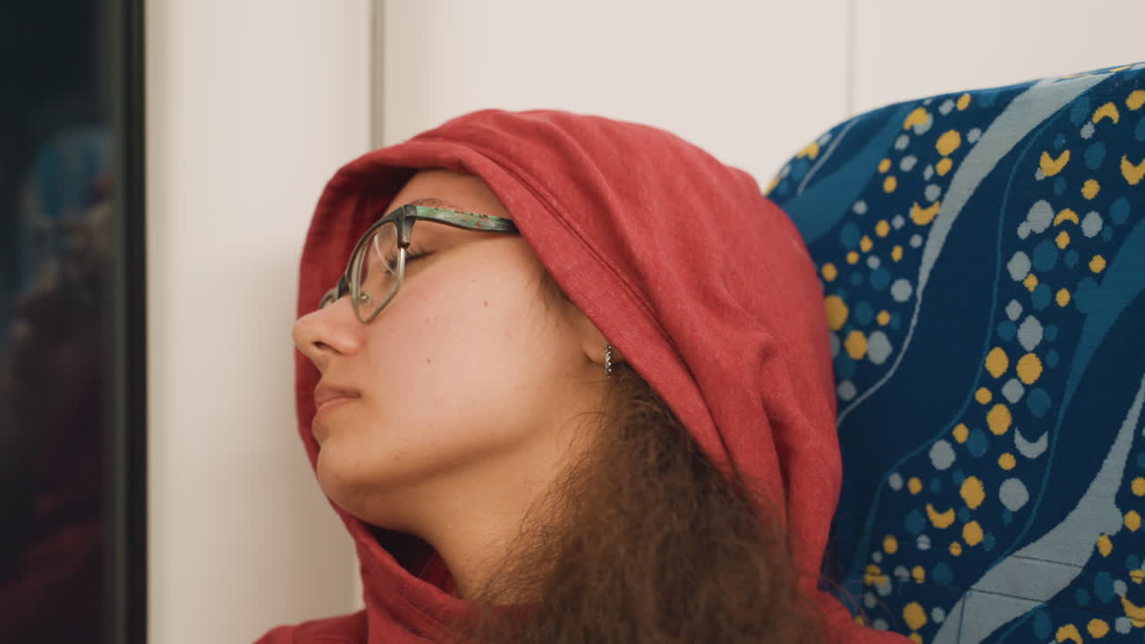 Penumpang yang tertidur di samping jendela kereta, kepala miring dan mata terpejam dalam jaket hoodie merah. Seorang pelancong larut malam melayang di antara stasiun, postur santai, cahaya gerbong yang lembut, siluet kota samar di luar, suasana sunyi.