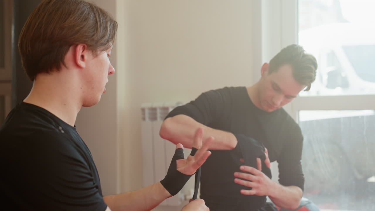 Two athletes wrap hands with black straps in training gym, preparing for boxing or martial arts practice with discipline and concentration, focusing on protection, readiness for workout