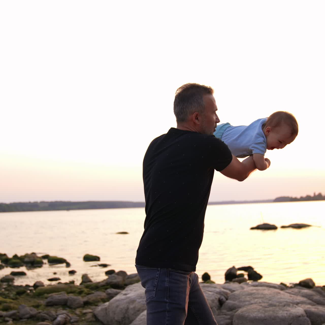 Un bellissimo neonato che vola tra le mani del suo premuroso papà. Una famiglia che trascorre del tempo sulla riva del fiume alla fine della giornata