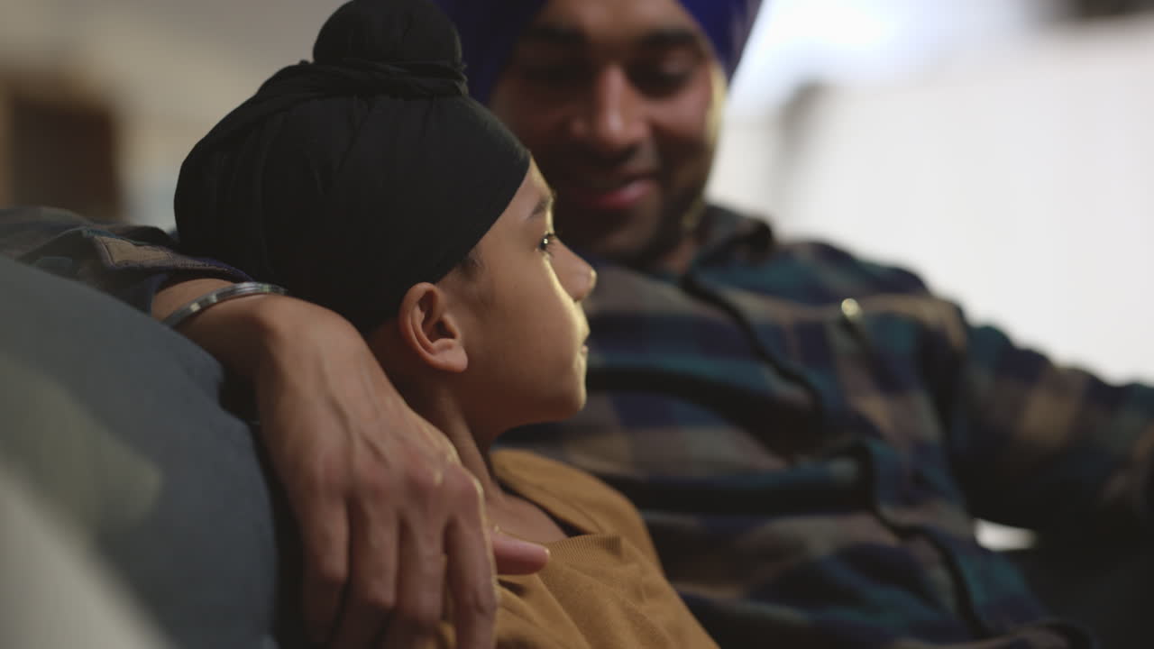 Close Up Of Loving Sikh Father And Son Wearing Turbans Sitting On Sofa At Home Talking Together Shot In Real Time