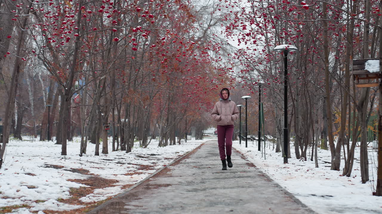 Jovem senhora com roupa de inverno e as mãos nos bolsos passeando por uma rua nevada e solitária, ladeada por árvores despidas e bagas vermelhas, em uma atmosfera fria com pegadas no caminho gelado e edifícios distantes.