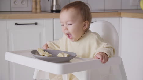 linda niña comiendo rodajas de plátano sentada en su trona en la cocina 1