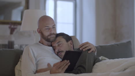 cute gay lying in bed with partner and reading news on tablet