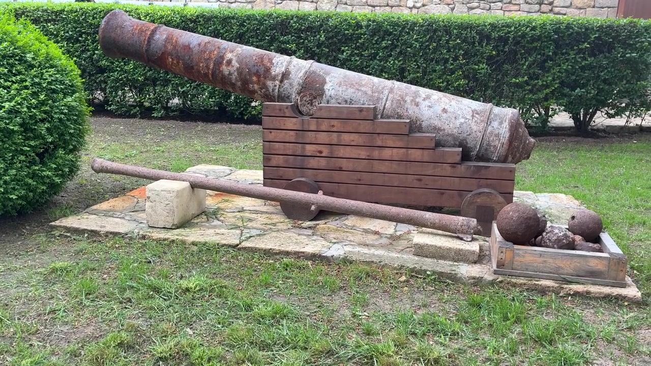 Um canhão antigo enferrujado sobre um reparo de madeira com balas de canhão, assente numa base de pedra numa área verde gramada, com uma sebe e uma muralha de fortaleza ao fundo.
