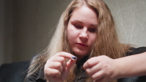 Close view seamstress seated on sofa, eyes down, focusing on threading needle with steady hands, blue nails visible, soft indoor light, patient expression, quiet repair moment in living room