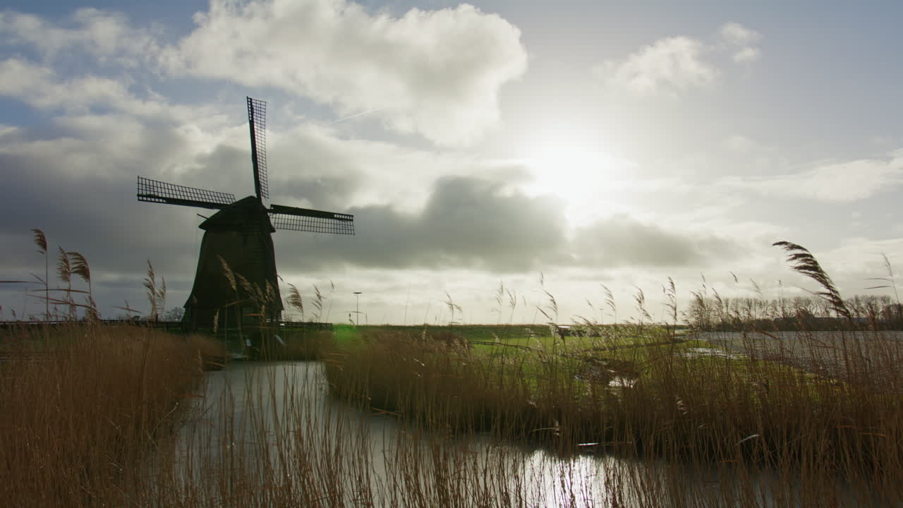 Wijd uitzicht op een traditionele windmolen in een open landschap met riet, water en een dramatische lucht, geschikt voor concepten als duurzame energie, traditie, duurzaamheid en rurale infrastructuur