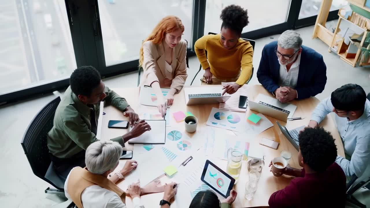 Top-down video view of diverse team collaborating around a table with charts and laptops