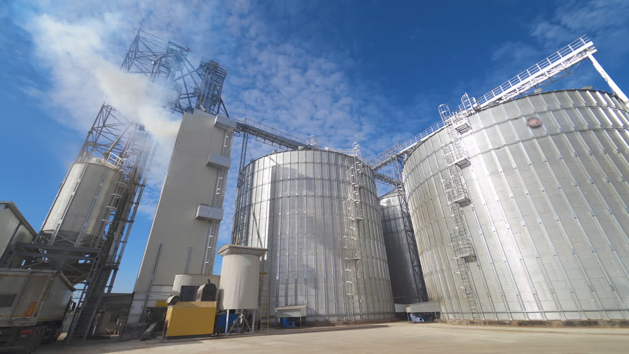 Huge hangar buildings. Modern grain elevators under blue sky. White vapor released from metal construction for drying grains. Panoramic view.