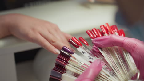 closeup of nail technician in pink gloves matching various red and purple nail polish tips on customer finger to help choose preferred shade and length during manicure consultation at salon
