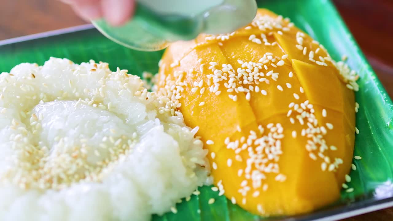 A close-up of coconut milk being poured over mango sticky rice, garnished with sesame seeds on a green plate.