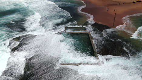 slow motion aerial shot of waves and powerful ocean swell crashing over monavale rock pool on a beautiful spring day