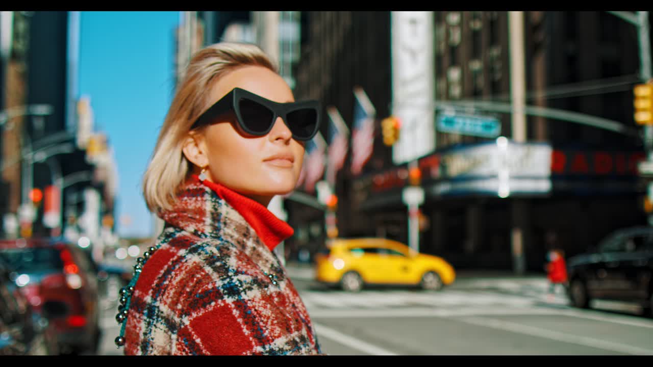 New York, USA, 9 ottobre 2025: Donna in cappotto a quadri e occhiali da sole vicino alla Radio City Music Hall. Donna bionda in cappotto a quadri e occhiali da sole neri in piedi su una strada di Manhattan con lo sfondo del teatro.