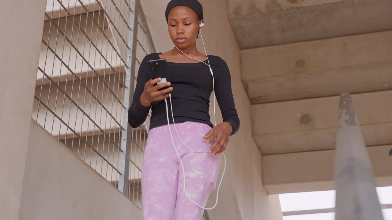 Donna di colore che prova un discorso su una scala, scorrendo il telefono per gli appunti mentre pratica i gesti, auricolari con filo, postura sicura e movimento espressivo della mano contro cemento grezzo e ambiente urbano.