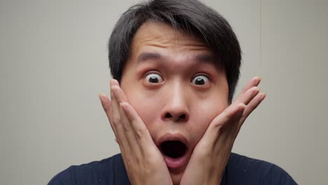 Closeup face shot of southeast asian man showing surprise opening eyes hands holding face, facial expression elevating eyebrows