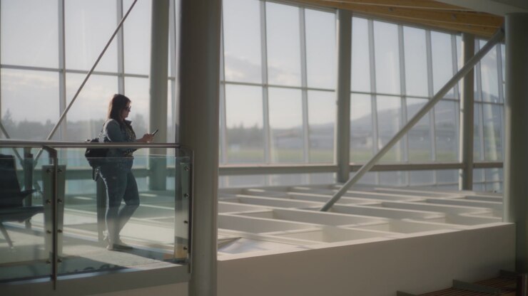 Mujer usando su teléfono mientras se apoya en una barandilla de cristal en la terminal del aeropuerto.