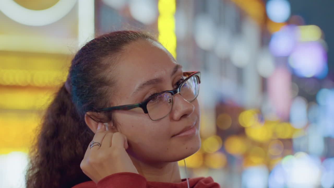 Giovane donna con auricolari, luci neon della città. Turista casual con felpa rossa inclina la testa e si aggiusta gli occhiali ascoltando musica. Bokeh vibrante di insegne luminose e bagliore del centro. Sorrisi spontanei e sguardo pensieroso.