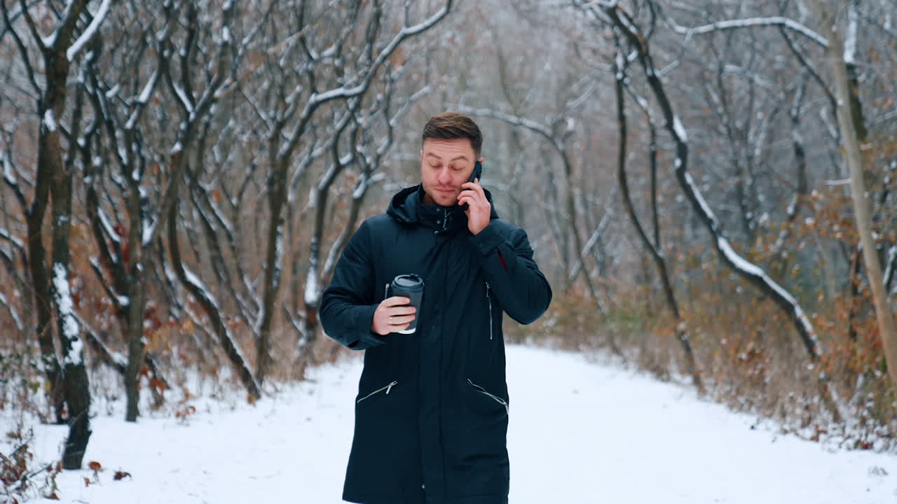 Man with coffee cup in hand speaks on the phone. Caucasian male walking by the beautiful park in winter.